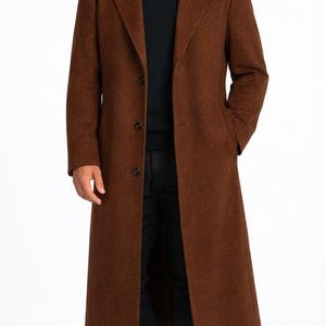 May include: A long, brown overcoat with a classic design. The coat features a notched lapel, three-button closure, and front pockets. The person is wearing a black shirt and black pants.