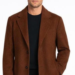 May include: A brown wool coat with a notched lapel and two-button closure. The coat is worn over a black crew neck shirt. The coat has a classic, tailored design.