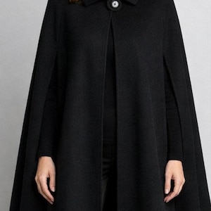 May include: A solid black cape featuring a structured collar and a single large circular button closure at the neckline. The garment has a flared silhouette with arm slits on the sides, worn over a black turtleneck sweater and black trousers.
