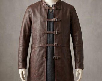 May include: A dark brown leather coat featuring a mandarin collar and four horizontal straps across the front, each secured with a silver-toned metal buckle. The coat is worn over a dark gray tunic with a small V-neckline, displayed on a mannequin.