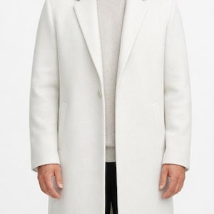 May include: A long, off-white overcoat featuring a notched lapel, a single-button closure, and two side pockets. The coat is worn over a light beige sweater and black trousers, creating a clean, structured silhouette with long sleeves.
