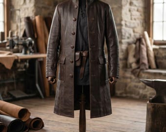 May include: A long, dark brown leather coat featuring a high collar, a front button closure with five visible buttons, and two flap pockets at the waist. The coat is displayed on a mannequin, layered over a black shirt with a small leather pouch.