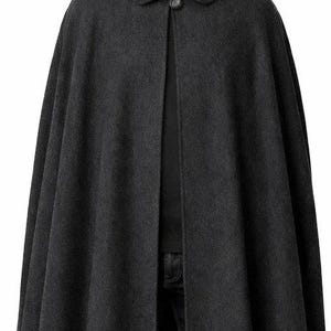 May include: A charcoal gray cape with a rounded collar and a single button closure. The cape is made of a thick, woven material and falls to the mid-thigh. The person in the image is wearing a black shirt and dark jeans.