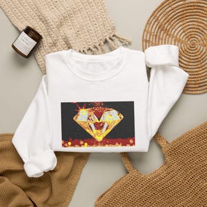May include: A white long-sleeve sweatshirt featuring a rectangular graphic print on the chest. The graphic depicts a large, faceted yellow diamond with red and orange accents against a dark background, with a red textured base at the bottom.