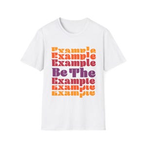 May include: White t-shirt featuring the words "Example" and "Be The Example" in a gradient of orange, red, and purple. The tee has a crew neck and short sleeves, suitable for casual wear.
