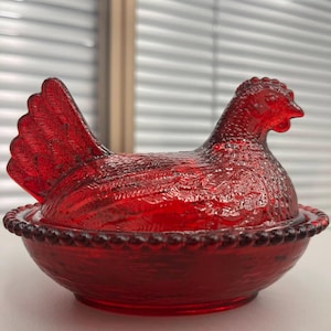 May include: A vibrant red glass hen-shaped dish with a textured surface. The hen's body forms a lid, and the base is a round bowl with a beaded rim. The design features detailed feather patterns, creating a decorative and functional piece.