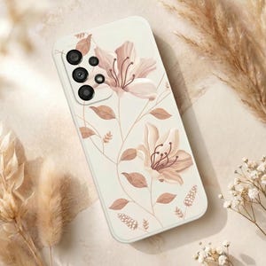 May include: A white phone case featuring a floral design with light pink and brown flowers and leaves. The case has a camera cutout and a smooth finish. The floral pattern is detailed and covers most of the case's surface.