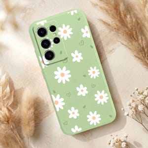 Puede incluir: Funda de teléfono verde claro adornada con margaritas blancas y pequeños corazones verdes. La funda tiene un acabado liso y está diseñada para un teléfono inteligente, con recortes para la cámara y los botones. El estampado floral añade un toque primaveral.