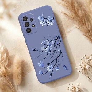 以下が含まれることがあります： 青い花と暗い枝のフローラルデザインが特徴の、ペリウィンクル色の携帯電話ケース。ケースはマット仕上げで、左上隅にカメラの切り抜きがあります。小さな蝶も確認できます。