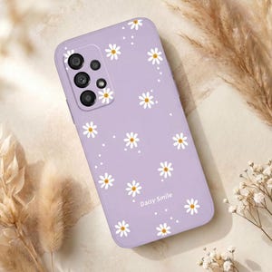 May include: A lavender phone case adorned with white daisies and small white dots. The case has a camera cutout and the words "Daisy Smile" are printed on the lower right. The case is designed to protect the phone.