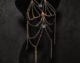 May include: A gold-toned body chain featuring multiple delicate draped chains and two clear butterfly-shaped charms. The jewellery piece includes a necklace section and extends down the torso with layered strands, creating a shimmering effect.