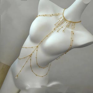 May include: Gold-toned body chain necklace with multiple strands and delicate beaded accents. The necklace is draped over a white mannequin, showcasing its design. The body jewelry features a choker-style neckline and cascading strands.