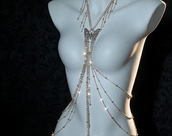 May include: A silver-toned body chain featuring a central butterfly pendant with intricate filigree detailing. The piece is constructed from multiple delicate link chains adorned with small metallic beads, creating a layered effect that drapes across the torso.
