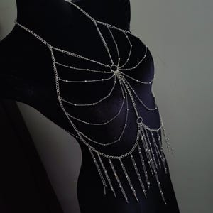May include: Silver body chain with multiple strands and delicate beaded accents. The chain drapes over the chest and torso, with dangling strands of beads. The jewelry is displayed on a black mannequin, showcasing its intricate design and elegant style.