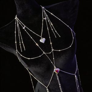 Pode incluir: Uma corrente corporal em tom prateado com múltiplas correntes de elos delicados e pequenas contas metálicas. O design inclui um pingente de coração prateado central e um coração inferior com um acabamento iridescente vermelho e roxo.
