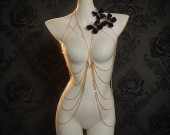 May include: A gold-toned body chain featuring multiple draped metallic strands that wrap around the torso. The accessory is accented with three black fabric butterfly embellishments positioned on the upper left side near the neckline.
