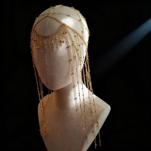 May include: A gold-toned headpiece featuring a delicate chain framework that rests on the head, adorned with multiple dangling chain strands and small metallic beads that drape down the sides, creating a layered and intricate ornamental accessory.