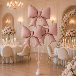 May include: Three large, matte dusty pink bow-shaped foil balloons are tethered together by thin pink ribbons to a small, weighted base. The balloons are arranged in a cluster, with one positioned above two others, creating a decorative display.