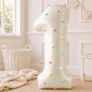 May include: A large, cream-colored foil balloon shaped like the number one, decorated with small, colorful pom-poms in shades of pink, blue, yellow, purple, and white attached to its surface. The balloon has a smooth, reflective finish.
