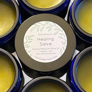 May include: Close-up of several small, round jars of healing salve. The salve is a yellow color and is surrounded by dark blue jars. The central jar has a label that reads "Healing Salve" with the ingredients and brand name.