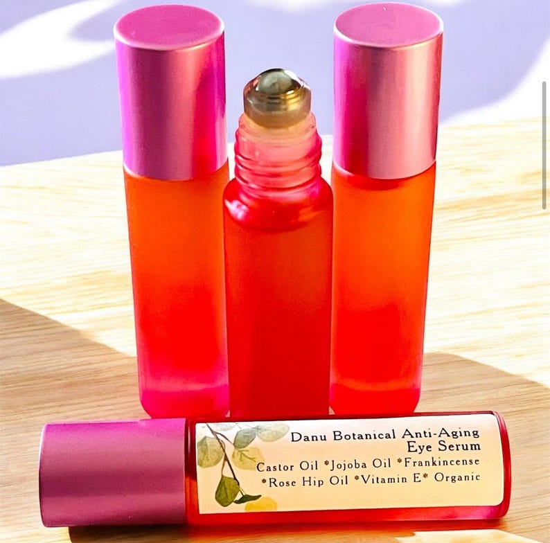 Puede incluir: Cuatro peque&ntilde;os frascos de vidrio naranja vibrante con tapas rosas y un aplicador roll-on. Una botella est&aacute; en horizontal, mostrando el texto "Danu Botanical Anti-Aging Eye Serum" con los ingredientes. Sobre una superficie de madera clara.