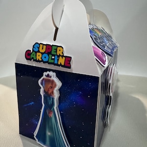 K&ouml;nnte beinhalten: Eine wei&szlig;e Geschenkbox mit Giebeldach und Griff, mit einem farbenfrohen "Super Caroline"-Logo. Die Box ist mit einer Prinzessinnen-Grafik und einem Weltraum-Hintergrund verziert. Die Box ist f&uuml;r Partygeschenke konzipiert.