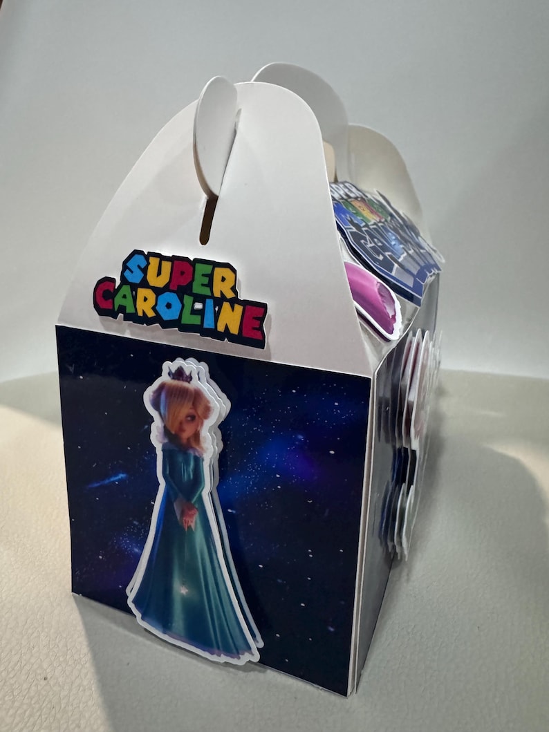 K&ouml;nnte beinhalten: Eine wei&szlig;e Geschenkbox mit Giebeldach und Griff, mit einem farbenfrohen "Super Caroline"-Logo. Die Box ist mit einer Prinzessinnen-Grafik und einem Weltraum-Hintergrund verziert. Die Box ist f&uuml;r Partygeschenke konzipiert.