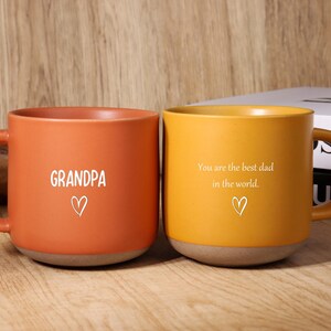May include: Two ceramic mugs, one orange and one yellow, with brown bases and handles. The orange mug has the word "GRANDPA" and a heart. The yellow mug has the text "You are the best dad in the world." and a heart.