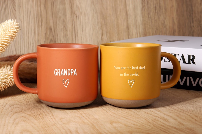 May include: Two ceramic mugs, one orange and one yellow, with brown bases and handles. The orange mug has the word "GRANDPA" and a heart. The yellow mug has the text "You are the best dad in the world." and a heart.