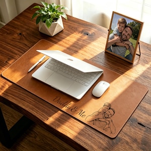 Könnte beinhalten: Eine braune Leder-Schreibtischunterlage mit dem Text "Daddy & Me" und einer Skizze eines Vaters und Kindes. Ein Laptop, ein weißer Stift und eine Maus liegen auf der Unterlage. Ein gerahmtes Foto von Vater und Kind steht auf dem Schreibtisch.