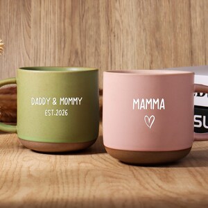 May include: Two ceramic mugs, one green and one pink, with brown bases and handles. The green mug says "Daddy & Mommy EST.2026" in white. The pink mug says "Mamma" with a heart symbol. Both mugs are on a wooden surface.