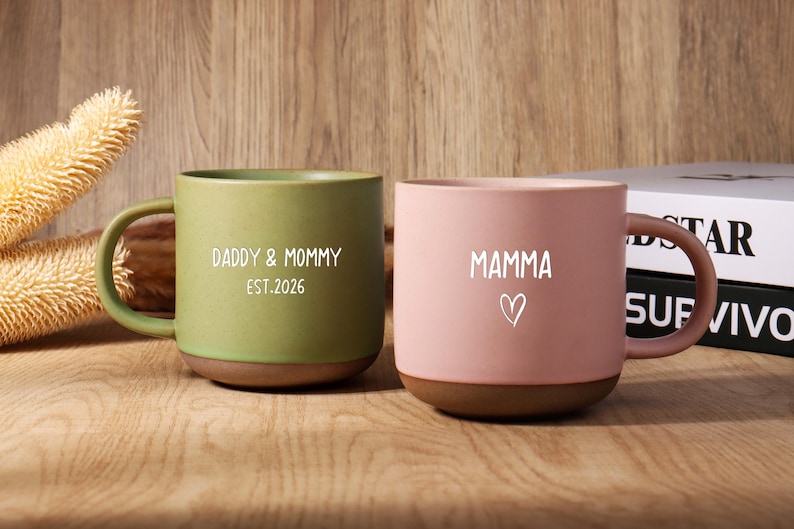 May include: Two ceramic mugs, one green and one pink, with brown bases and handles. The green mug says "Daddy & Mommy EST.2026" in white. The pink mug says "Mamma" with a heart symbol. Both mugs are on a wooden surface.