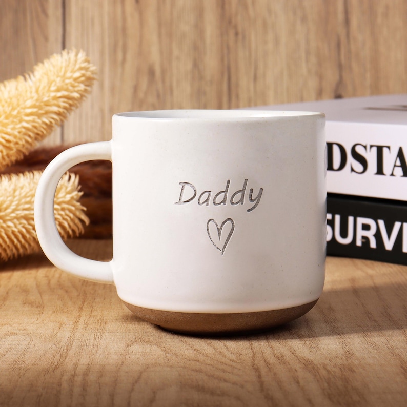 May include: White ceramic mug with a brown base and a large handle. The word "Daddy" and a heart are etched on the front. The mug is on a wooden surface.