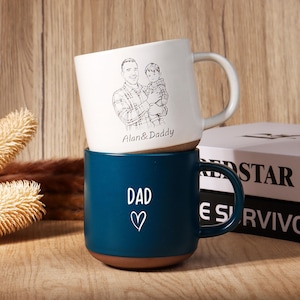 May include: Two ceramic mugs stacked on top of each other. The top mug is white with a black sketch of a man holding a child, with the text "Alan & Daddy". The bottom mug is teal with the word "DAD" and a heart.