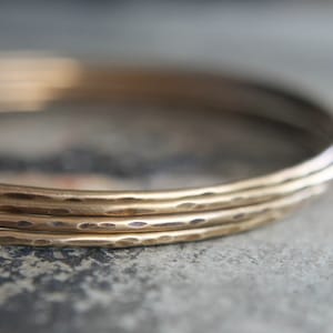 May include: Close-up of several gold-colored bangles. The bangles have a hammered texture, giving them a unique, handcrafted appearance. The jewelry is stacked, creating a layered effect. The background is a blurred, neutral tone.
