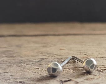 May include: A pair of small, silver stud earrings with a textured, hammered finish. The earrings are round and sit on a wooden surface with a dark background. The studs are simple and minimalist.