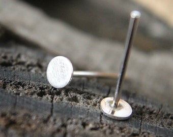 May include: Close-up of two silver labret studs. Each stud features a small, flat, circular disc attached to a straight post. The studs are displayed on a weathered, dark wood surface, highlighting their minimalist design.