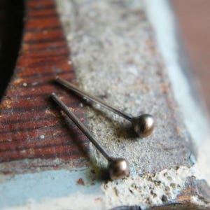 May include: Two small, antique-looking metal pins with a ball-shaped head. The pins are a bronze color and are lying on a weathered, multi-colored surface. The pins are likely used for jewelry making or crafting.