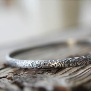 May include: A thin, circular silver bangle bracelet featuring a detailed, embossed floral and vine pattern that wraps around the entire exterior surface. The metal has a darkened, oxidized finish within the recessed areas to highlight the design.