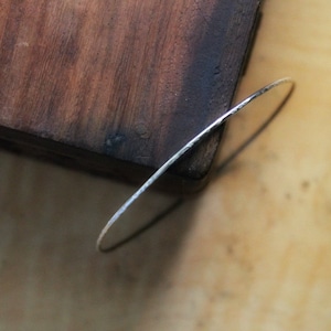 Peut inclure: Un fin bracelet jonc en argent avec une texture martelée. Le bracelet repose sur une surface en bois, mettant en valeur son design délicat et son savoir-faire. La forme circulaire du jonc est mise en évidence.