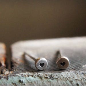 May include: A pair of silver stud earrings. Each earring features a small, round design with a heart-shaped cutout in the center. The earrings are set against a blurred, neutral background.