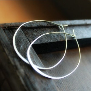 May include: A pair of silver-coloured hoop earrings with a teardrop design. The earrings are displayed on a dark wooden surface. The thin, minimalist hoops reflect light, creating a subtle shimmer.