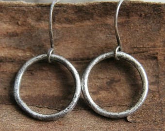 May include: A pair of silver hoop earrings. The earrings are circular with a textured surface and a simple hook closure. The earrings are displayed on a wooden surface, showcasing their handcrafted appearance. The earrings are a classic jewelry item.