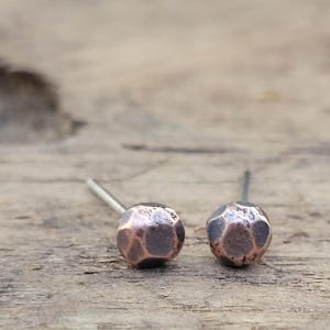 May include: A pair of copper-colored stud earrings. Each earring features a textured, faceted design. The earrings have a post backing for pierced ears. The earrings are displayed on a wooden surface.
