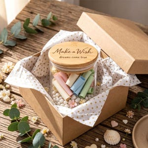 Pode incluir: Um frasco de vidro transparente com tampa de madeira com o texto Make a Wish dreams come true, preenchido com pequenos rolos de papel em tons pastel e contas brancas, dentro de uma caixa de presente de papelão castanho com papel de seda dourado.