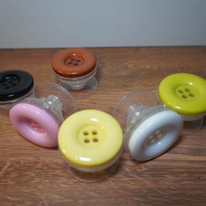 May include: A collection of six circular phone grips, each designed to resemble a large button with four central holes. The grips feature glossy tops in black, brown, pink, yellow, white, and lime green, all attached to clear plastic bases.