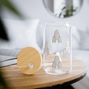 Op de afbeelding: Een helder glazen drinkglas in de vorm van een blikje met een bamboe deksel en rietje. Het glas is versierd met een herhalend patroon van kleine, spookachtige figuren. Het deksel en de tafel zijn van hout.