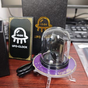 May include: A UFO-Clock with a clear dome and a purple base, featuring the text "UFO-CLOCK" and "10:24". The clock sits on clear legs and is accompanied by a black box with a gold UFO logo.