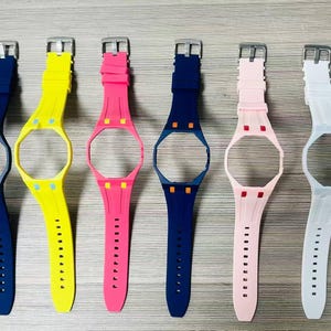 May include: A collection of eight silicone watch bands with integrated protective cases, arranged in a row. The bands are available in black, navy blue, yellow, hot pink, dark blue, light pink, white, and bright green, each with a metal buckle.