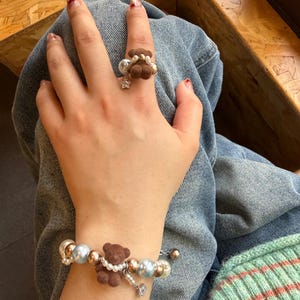 May include: A brown teddy bear ring and bracelet set. The ring features a small brown bear with pearl accents and a star-shaped charm. The bracelet includes a brown bear, pearls, and gold-colored beads. The jewelry is set against a denim background.